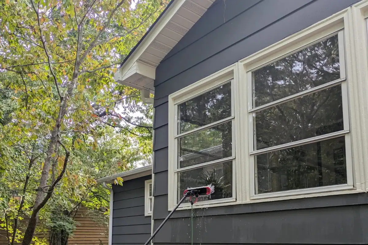 A long-handled brush sprays water while cleaning the exterior windows of a dark gray house, surrounded by trees with green leaves.
