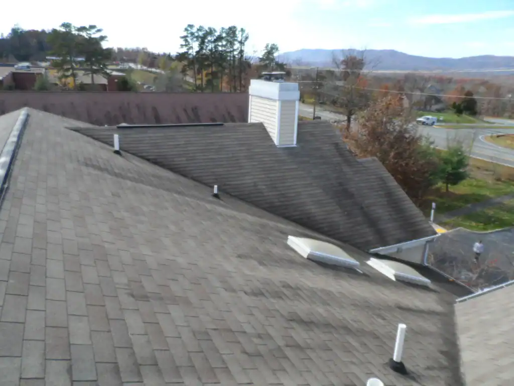 View of a sloped asphalt shingle roof with several vent pipes, skylights, and a white rooftop structure. Trees, a road, and distant hills are visible in the background under a blue sky.