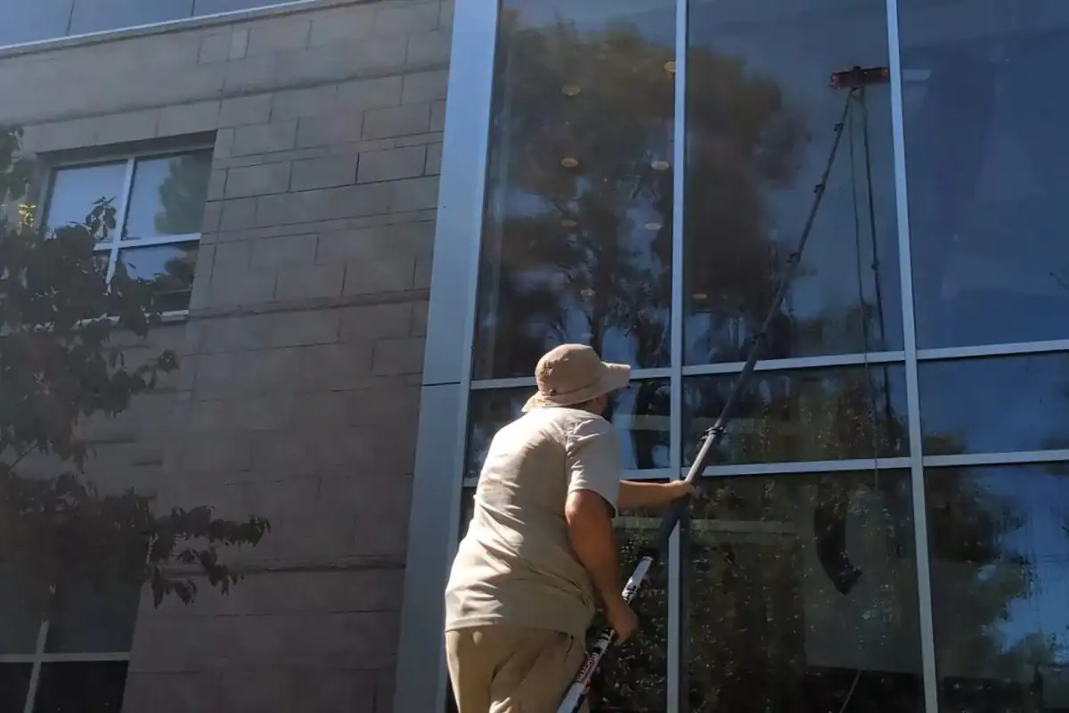 A person wearing a hat and light-colored clothing uses a long pole to clean the tall glass windows of a building on a sunny day.