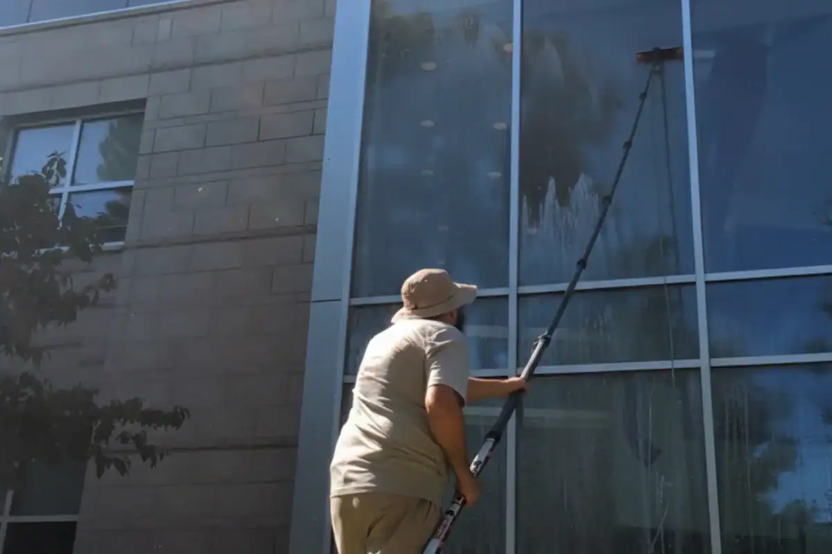 A person wearing a hat and beige clothing uses an extension pole to wash the large glass windows of a building from the ground.