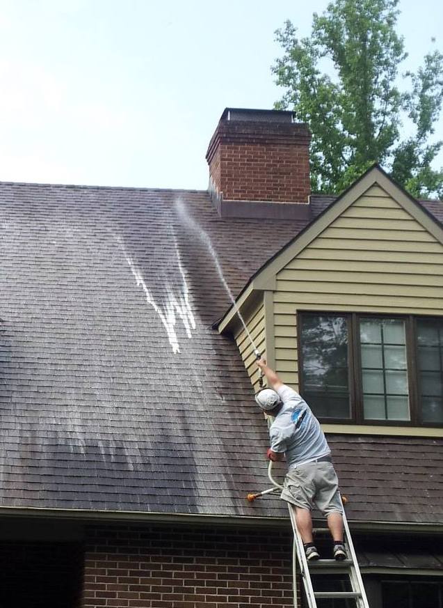 A person standing on a ladder is cleaning a house roof with a pressure washer, demonstrating how to remove algae from roof surfaces. Water sprays away dirt and debris, leaving visible clean streaks near a large brick chimney.